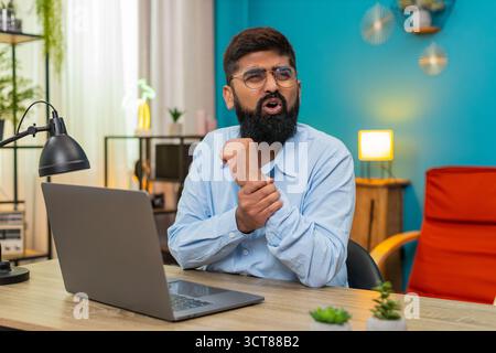 Indischer Mann zu Hause massiert Wunde Handgelenke nach langer Laptop-Sitzung Schmerzen von wiederholter Belastung. Ein Araber am Schreibtisch beugt die Finger und spürt, dass der Schmerz lindert, indem er sanfte Übungen durchführt, in der Hoffnung, dass sich die Sehnen erholen. Stockfoto