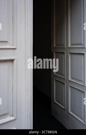 Teilweise offene weiße Holztür führt in dunkle Innenräume und symbolisiert Mysterium, Gelegenheit und Übergang Stockfoto
