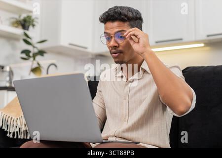 Ein indischer Mann zu Hause hat Schwierigkeiten, winzige Texte auf dem Laptop zu lesen, zieht dann eine Brille auf und sieht klare Sicht. Arabian Guy on Sofa Lächeln passt den Rahmen an, liest Bildschirmlinien ohne Belastung und setzt die Arbeit fort. Stockfoto