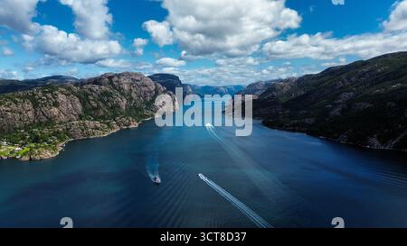 Aus der Vogelperspektive auf den norwegischen Fjord Lysefjord mit Booten, die durch klares blaues Wasser segeln, umgeben von steilen Bergklippen unter einem hellen Sommerhimmel Stockfoto