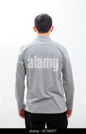 Ein Mann steht mit dem Rücken zur Kamera, trägt ein sauberes graues, langärmeliges Poloshirt und eine schwarze Hose vor einem nahtlosen weißen Hintergrund. Stockfoto