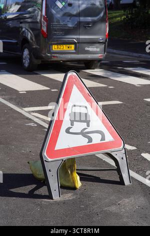 Vorübergehendes britisches Verkehrszeichen, das vor rutschigen Bedingungen warnt, auf einer städtischen Straße mit einem Fahrzeug und einer Zebraüberquerung im Hintergrund angebracht wird. Dorking Stockfoto