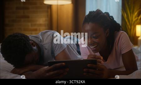 Fröhlich lachend afroamerikanisch verheiratetes Paar zwei Leute Familie Ehefrau Ehemann liegt auf dem Bett und hält ein digitales Tablet. Sehen Sie sich lustige Video-Film-Komödie an Stockfoto