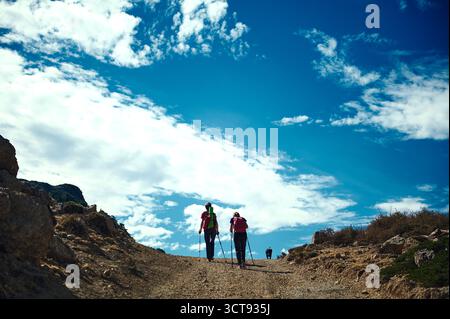 Zwei Wanderer mit Rucksäcken wandern einen felsigen Pfad entlang eines Hügels unter einem klaren blauen Himmel. Ein lebendiger Outdoor-Moment für Abenteuer, Fitness und gemeinsame Nutzung Stockfoto