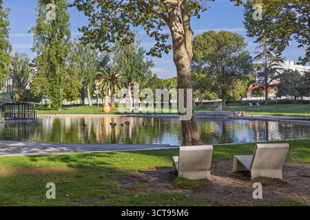 Eine friedliche Szene im Parque do Bonfim, Setúbal, mit weißen, eckigen Bänken im Schatten eines großen Baumes mit Blick auf den zentralen See und die Enten. Stockfoto