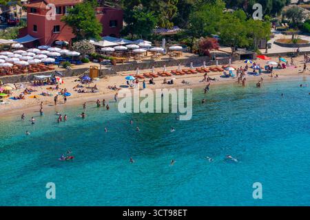 Aus der Vogelperspektive auf einen atemberaubenden überfüllten Strand in Griechenland mit türkisfarbenem Wasser, einem Restaurant am Meer und Reihen weißer Sonnenschirme an einem perfekten Sommertag Stockfoto