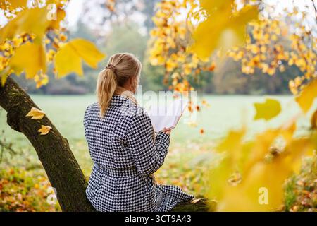Frau im Hahnentrittmantel sitzt am Baum und liest Buch im Herbstpark. Entspannung im Freien Stockfoto