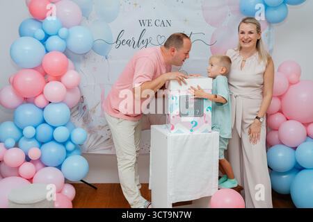 Vater und Sohn schauen sich die dekorierte Box an, lächelnde Mutter in der Nähe während der Babyparty. Herzlicher Familienmoment für Event-Promotion, Elternschaft Stockfoto