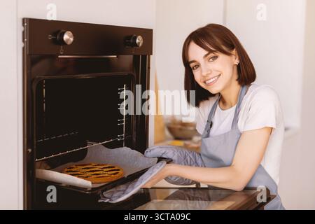 Porträt einer glücklichen Dame, die in der Küche backt, Schürze und Fäustling trägt und das Blech mit hausgemachtem Kuchen aus dem heißen Ofen nimmt Stockfoto