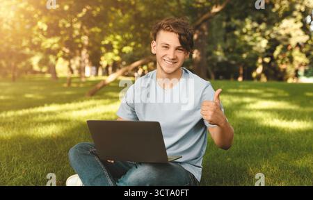 Glücklicher männlicher Student, der wie gestikuliert, das Online-Bildungsprogramm genehmigt, mit einem Laptop im Park sitzt und den Daumen nach oben zeigt Stockfoto