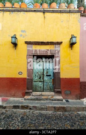 Eine verwitterte, farbenfrohe Fassade in San Miguel de Allende, Mexiko, mit einer gelben Wand mit rotem Sockel, einer rustikalen blauen Tür, die von Stein eingerahmt und vergossen ist Stockfoto