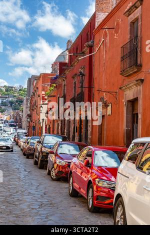 Autos parken entlang einer Kopfsteinpflasterstraße vor roten Gebäuden in San Miguel de Allende, Mexiko. Stockfoto