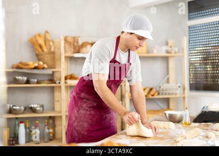 Porträt eines jungen Bäckers in Schürze, der mit Teig arbeitet, Bäckereiprodukte kneten und Formen in der Küche in der Bäckerei Stockfoto