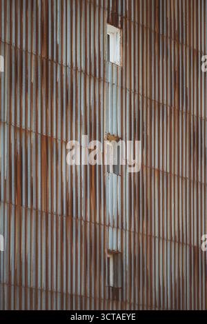 Vertikale Tele-Ansicht einer industriellen Metallwand mit rostiger Wellpappe und kleinen quadratischen Fenstern, mit veralteten Stahlflächen und geomet Stockfoto