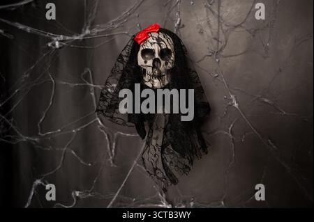 Ein gruseliger dekorativer Schädel mit schwarzem Spitzenschleier und einer leuchtend roten Blume vor dunklem Spinnennetz Hintergrund. Halloween-Feiertagskonzept. Stockfoto