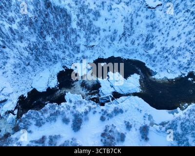 Aus nächster Nähe betrachten Sie einen dunklen Fluss mit starker Strömung und schäumenden Stromschnellen, umgeben von Eisbrocken und Neuschnee Stockfoto
