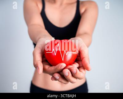 Rotes Herz mit Heartbeat Line Symbol in den Händen der Sportfrau, repräsentiert Cardio-Workout, Herzgesundheit und Fitness-Motivation. Das Konzept der körperlichen Aktivi Stockfoto