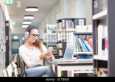 Junge Studentinnen, die in einer Bibliothek studieren. Sie sitzt an einem Schreibtisch, trägt Kopfhörer und benutzt ein Smartphone. Bildungskonzept, wissen Stockfoto