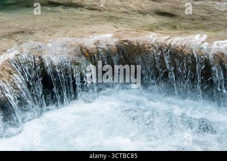 Nahaufnahme eines kleinen Wasserfalls, der über felsige Kanten mit klarem blauem Wasser und sanften Blasen fließt. Stockfoto