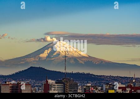 Ein Berg mit Schnee liegt im Hintergrund einer Stadt. Der Himmel ist blau und die Sonne untergeht. Panoramablick auf Mexiko-Stadt, schneebedeckten Vulkan Stockfoto