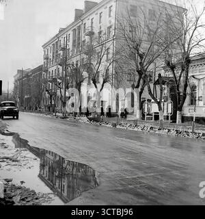 Ein stimmungsvoller Blick auf die Schewtschenko-Straße in Sloviansk, ukrainische SSR, während eines Wintertauens in den 1950er Jahren Die nasse Straße ist voller Pfützen, die stimmungsvolle Reflektionen der großen Stalinka-Gebäude erzeugen, die die Straße säumen. Ein klassischer GAZ-M20 Pobeda-Wagen, ein Wahrzeichen der Sowjetzeit der Nachkriegszeit, steht auf der Straße. Fußgänger versammeln sich auf dem Bürgersteig, warten oder gehen vorbei an der beeindruckenden neoklassizistischen Architektur Stalins. Diese ergreifende Szene fängt die düstere Schönheit der Nebensaison in einer Stadt ein, die sich nach dem Krieg erholt und wieder aufgebaut hat Stockfoto
