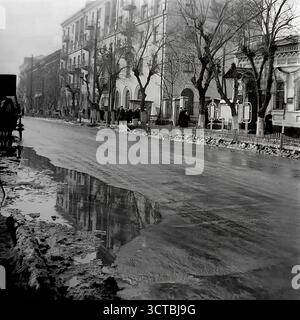 Ein stimmungsvoller Blick auf die Schewtschenko-Straße in Sloviansk, ukrainische SSR, während eines Wintertauens in den 1950er Jahren Die nasse Straße ist voller Pfützen, die stimmungsvolle Reflektionen der großen Stalinka-Gebäude erzeugen, die die Straße säumen. Ein klassischer GAZ-M20 Pobeda-Wagen, ein Wahrzeichen der Sowjetzeit der Nachkriegszeit, steht auf der Straße. Fußgänger versammeln sich auf dem Bürgersteig, warten oder gehen vorbei an der beeindruckenden neoklassizistischen Architektur Stalins. Diese ergreifende Szene fängt die düstere Schönheit der Nebensaison in einer Stadt ein, die sich nach dem Krieg erholt und wieder aufgebaut hat Stockfoto