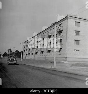 Ein neu fertiggestelltes Wohnviertel in der Chubarya-Straße (heute Svetlodarska-Straße) in den 1950er Jahren Sloviansk, ukrainische SSR. Die Fotos zeigen die Übergangsarchitektur der Epoche mit soliden, dreistöckigen Backsteinbauten, die vor dem „Chruschtschjowka“-Boom entstanden sind. Ein klassischer GAZ-M20 Pobeda-Wagen, ein Wahrzeichen der Nachkriegszeit, fährt auf der unbefestigten Feldstraße. Die Szene hebt den Kontrast der Zeit hervor: Brandneue, hochwertige Wohnungen wurden fertiggestellt, während die umliegende Infrastruktur, wie Straßen, noch in Entwicklung war. Stockfoto