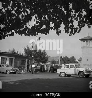 Eine belebte Tankstelle (AZS) in Sloviansk, ukrainische SSR, Ende der 1950er Jahre Dieses Archivfoto zeigt eine typische Szene aus der frühen Ära der sowjetischen Automobilisierung. Ein klassischer GAZ-M20 Pobeda und ein neuerer Moskvitch-407, legendäre Autos dieser Zeit, werden beim Tanken gesehen. Die Vintage-Pumps im Säulenstil und das Warnschild „Rauchen ist gefährlich!“ Tragen Sie zur authentischen Atmosphäre bei. Dieses Bild eines Schlüsselstücks der städtischen Infrastruktur ist ein mächtiges Symbol für die Modernisierung der Stadt nach dem Krieg und die zunehmende Mobilität ihrer Bevölkerung Stockfoto