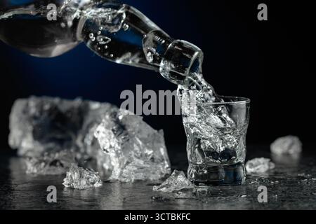 Kristallklarer Wodka oder Gin wird in ein Glas gegossen. Transparentes alkoholisches Getränk mit Eisstücken auf schwarzem Hintergrund. Stockfoto