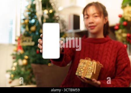 Weihnachtseinkäufe. Junge Frau, die Smartphone und Geschenkbox in festlicher Umgebung hält. Stockfoto
