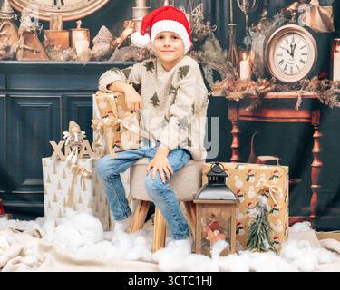 Junge mit Weihnachtsmann-Hut sitzt zwischen verpackten Paketen und festlicher Dekoration und hält ein kleines Geschenk. Gemütliche Urlaubsszene für saisonale Kataloge, Grußkarten und Stockfoto