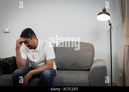 Ein nachdenklicher asiatischer Mann, Mitte 40, sitzt allein auf seiner Couch in einem schwach beleuchteten Raum und hält den Kopf tief in Gedanken. Dieses Bild stellt männliche mentale Heilung dar Stockfoto