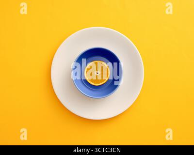 Die orangefarbene Scheibe liegt in einer blauen Schüssel, zentriert auf einer weißen Platte, wodurch eine minimalistische Komposition auf gelbem Hintergrund entsteht. Gesunde Ernährung oder Diät c Stockfoto
