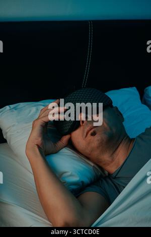 Ein Mann in einem Schlafzimmer hebt seine Schlafmaske mit einer Hand an, während er auf dem Rücken liegt, Kopf auf einem Kissen, ein weißes Bettlaken an seiner Brust gezogen, grau Stockfoto