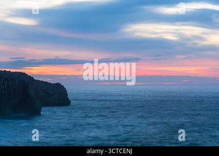 Sonnenuntergang über der felsigen Küste, Wellen krachen unter dem glühenden Horizont. Stockfoto