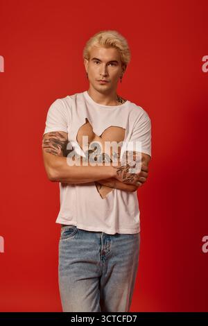 Junger Mann mit Tätowierungen steht selbstbewusst und zeigt jugendliche Mode in einem kühnen Setting. Stockfoto