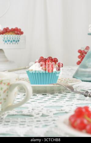 Cremefarbene Cupcakes mit roten Früchten auf Retro-Servietten, blaue Cupcake-Dosen, Vintage-Tasse und historische Gabel. Elegante Tischdekoration. Stockfoto