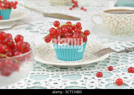Ein cremefarbener Cupcake mit roten Früchten; rote und blaue Tischdekoration: Tasse Kaffee oder Tee mit Milch auf Häkeltischdecke. Stockfoto