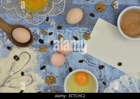 Backzubereitungen, viele Zutaten und ein leeres Stück Papier, auf das das Rezept geschrieben werden kann. Tisch mit Blick von oben und gestickte Retro-Servietten. Stockfoto