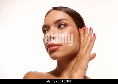 Eine junge Frau mit weichem Make-up berührt sanft ihr Gesicht vor einem weißen Hintergrund. Stockfoto
