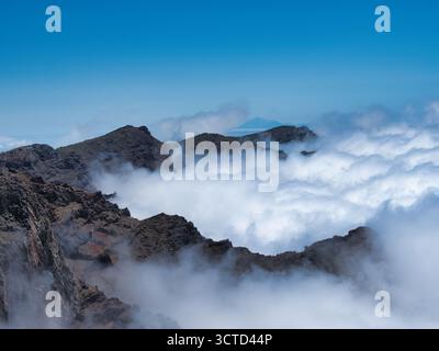 Volcanic Peaks und FAR Island Above Clouds - dramatische Bergsicht auf scharfe vulkanische Grate, die aus einem dichten Wolkenmeer hervorgehen, mit einem entfernten i Stockfoto