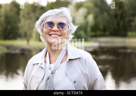 Glückliche Seniorin mit weißem Haar, die draußen lächelt, Sonnenbrille und Jacke trägt, einen aktiven Lebensstil im Ruhestand genießt, positives Altern, Freiheit, Freude Stockfoto