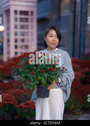 Schöne junge asiatische Frau, Käuferin oder Verkäufer, die einen Topf mit leuchtenden roten Chrysanthemen in der Hand hält. Urbane Herbstszene in der Nähe eines Blumenladens mit einem Rosa Stockfoto