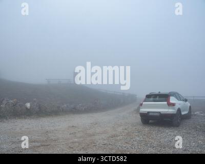 Weißer SUV parkt auf einer nebeligen Schotterstraße – weißer SUV parkt auf einer Schotterstraße in dickem, dichtem Nebel, mit einer schwachen Steinstruktur, die im dist kaum zu sehen ist Stockfoto