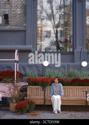 Stilvolle junge asiatische Frau, die auf einer Holzbank vor einem großen Blumenladen-Fenster sitzt. Die urbane Herbstszene zeigt leuchtende rote Chrysanthemen Stockfoto