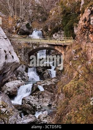 Steinbrücke und Wasserfall in der Mountain Gorge - vertikale Aufnahme einer rustikalen Steinbogenbrücke über einen kaskadierenden Fluss und Wasserfall inmitten der großen kuppe Stockfoto