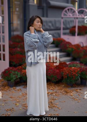 Nachdenkliche junge asiatische Frau, die zwischen gefallenen Blättern steht und ihren Pullover anpasst. Der Hintergrund ist mit leuchtenden roten Chrysanthemen und rosafarbenem Dekor versehen Stockfoto