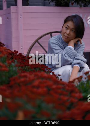 Junge asiatische Verkäuferin, die auf dem Boden zwischen leuchtenden roten Chrysanthemen-Blumen im Blumenladen sitzt. Sie blickt weg, neben einer rosa Holzblume Stockfoto