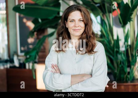 Eine schöne, selbstbewusste Frau steht mit überkreuzten Armen in einem modernen öffentlichen Raum und schaut in die Kamera. Stockfoto
