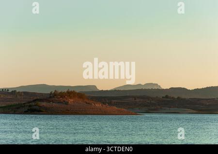 Ruhiges Seewasser, das das goldene Licht der untergehenden Sonne reflektiert, mit einem kleinen Hügel im Vordergrund und silhouettenreichen Bergen in der Ferne Stockfoto